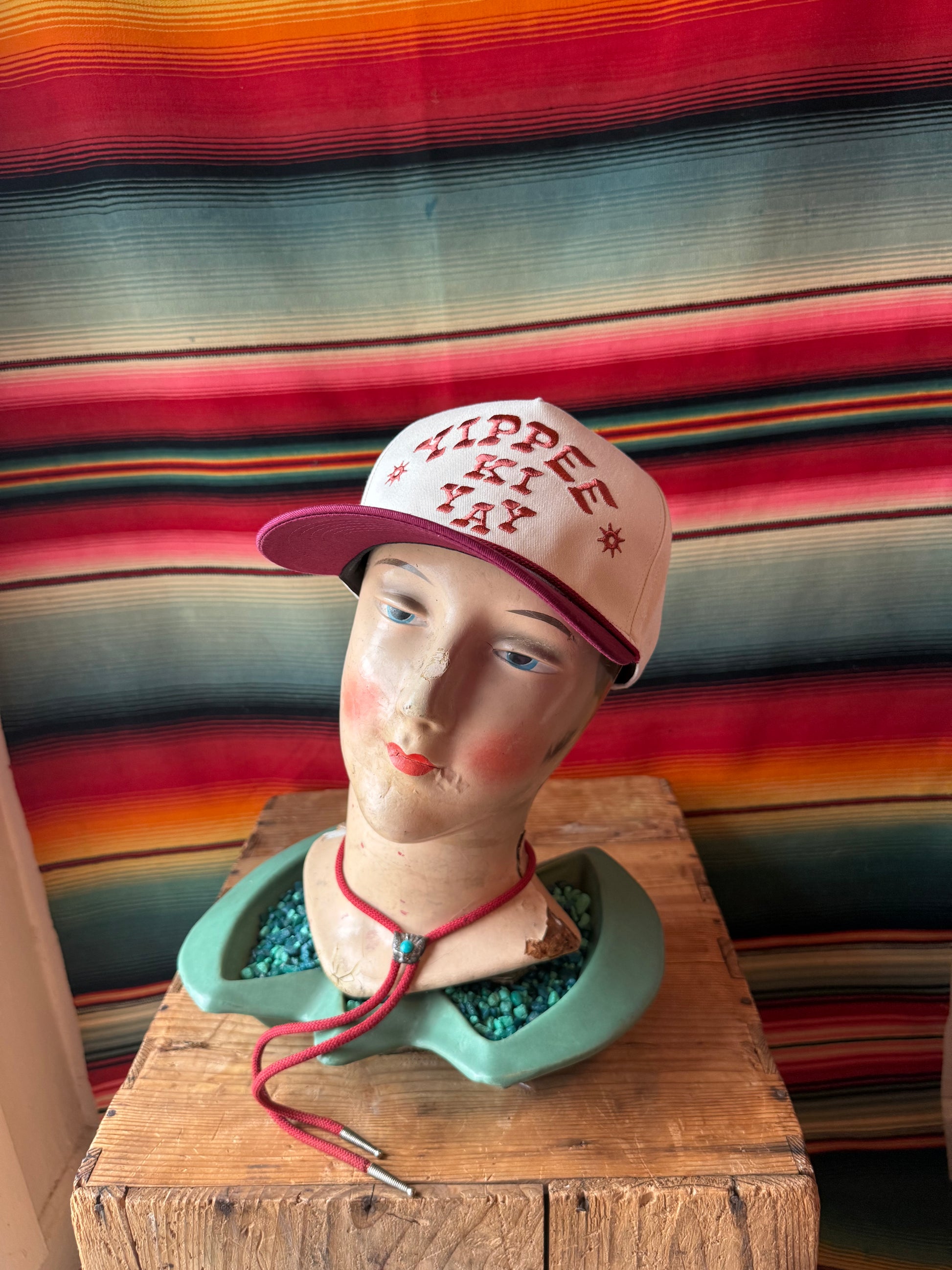 Mannequin head wearing a pink cap with 'Yippee Ki Yay' text against a colorful striped background.
